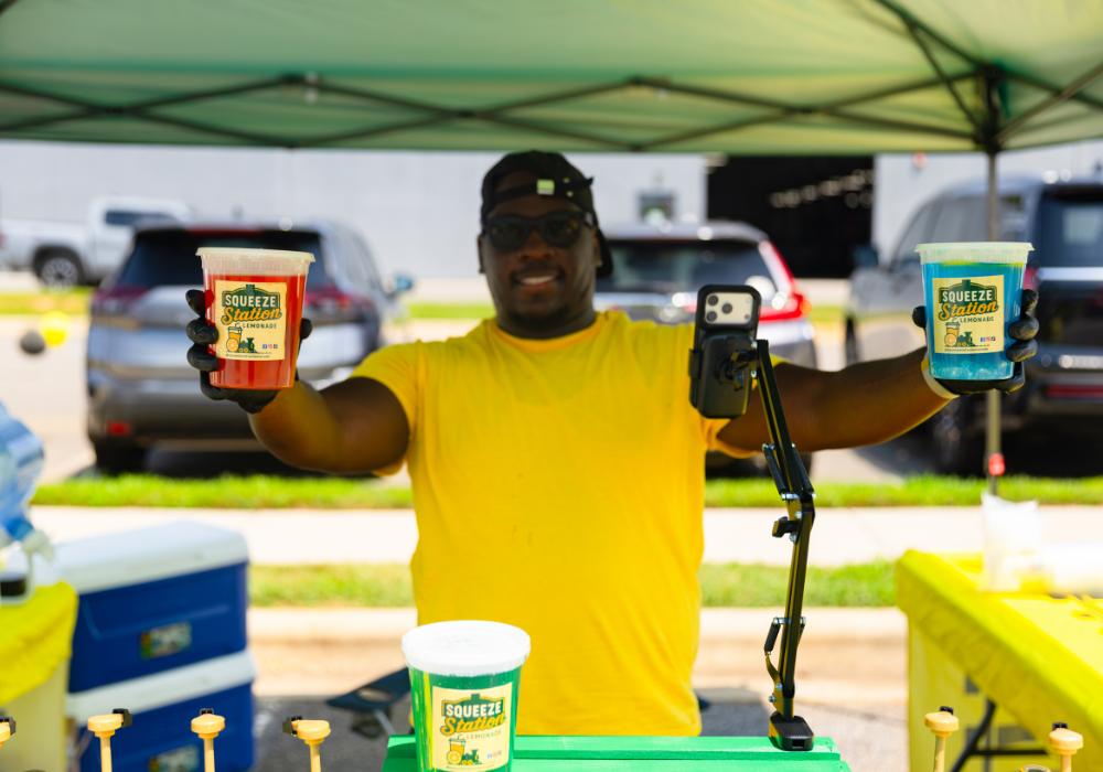 A man under a tent holding up two cups of lemonade